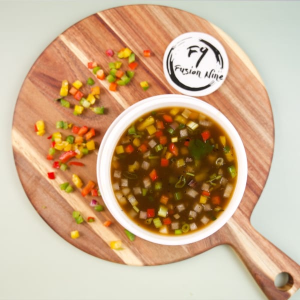 A bowl of dark soup with colorful diced vegetables on a wooden board with a Fusion Nine logo.