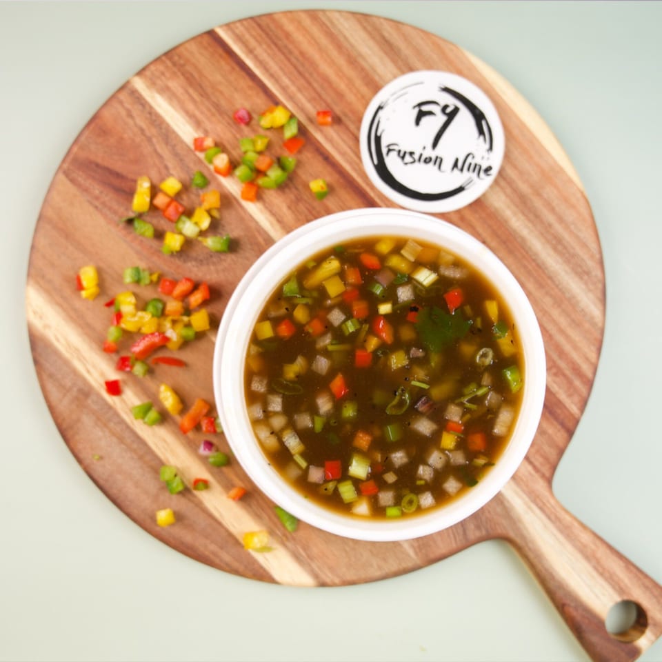 A bowl of dark soup with colorful diced vegetables on a wooden board with a Fusion Nine logo.