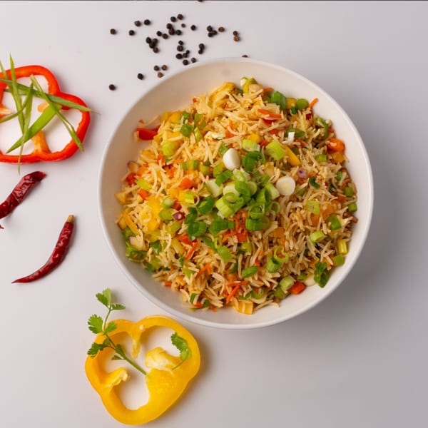 A white bowl of vegetable fried rice with bell peppers, chilies, and peppercorns on a white surface.