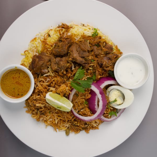 Mutton biryani with rice, red onions, lime, hard-boiled egg, and two sauces on a white plate.