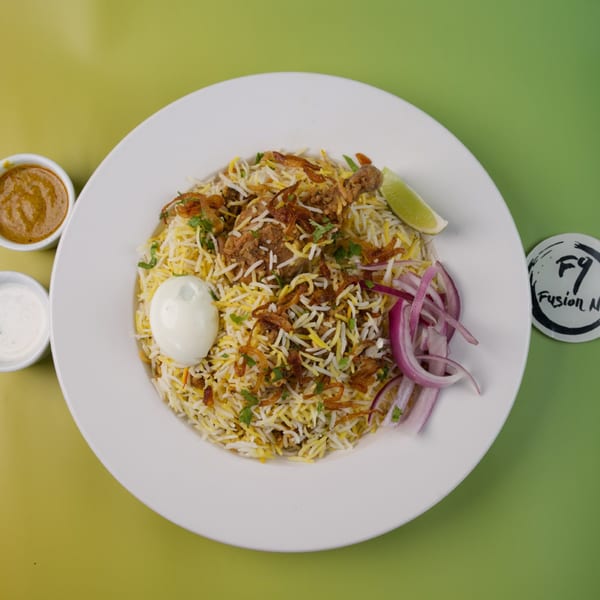 Chicken biryani with a boiled egg, red onions, lime, and two sauces on a white plate.
