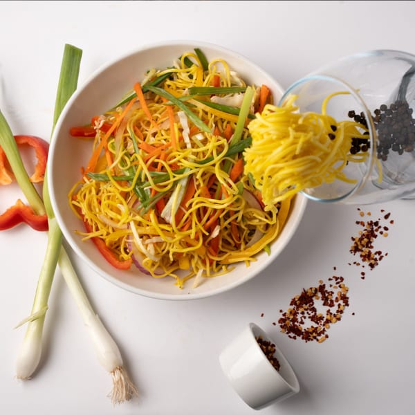 Noodles with vegetables in a white bowl, with spices and green onions on a white surface.