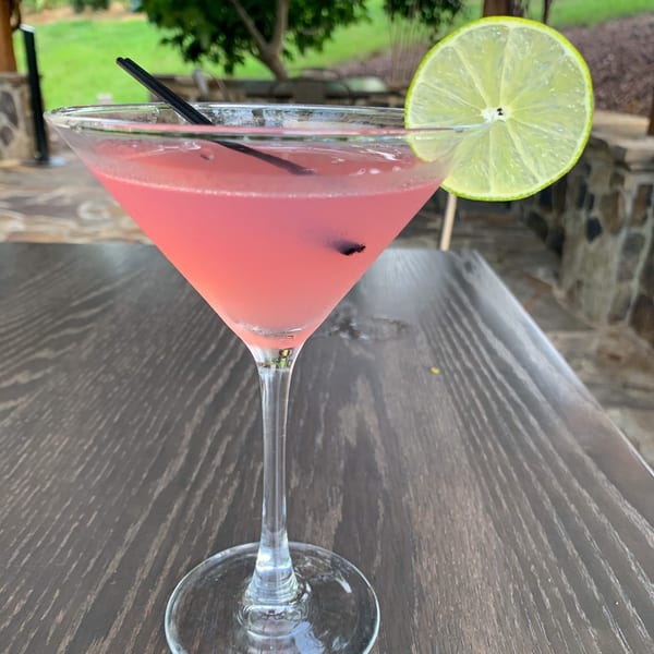 Pink cocktail in a martini glass with a lime slice and black straws on a wooden table.