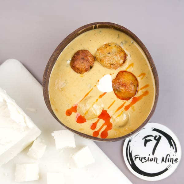 Overhead view of a coconut bowl filled with creamy curry, kofta balls, and red sauce, next to paneer.