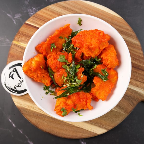 Bright orange fried chicken pieces, garnished with green herbs, in a white bowl on a wooden board.
