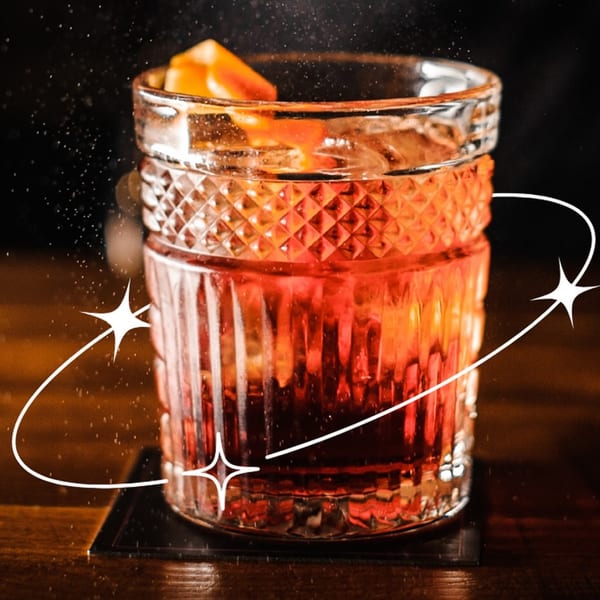 A vibrant red cocktail in a textured glass, garnished with orange, on a dark wooden surface.