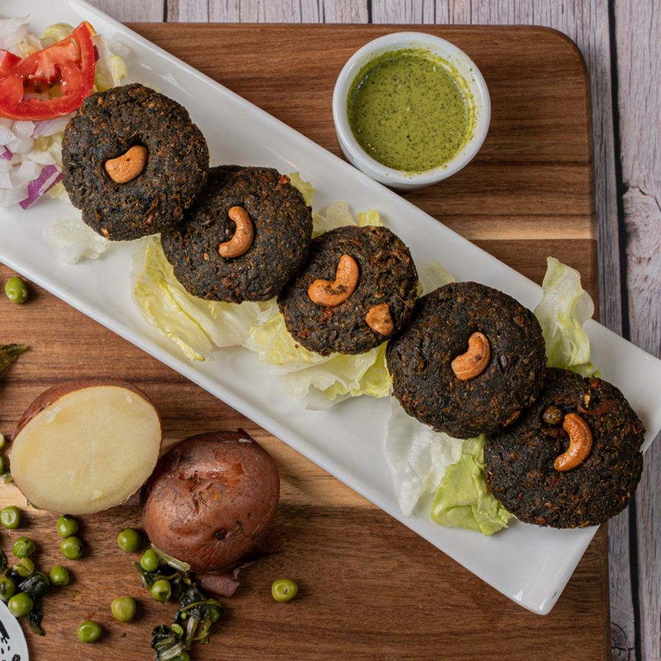 Dark brown patties with cashews on lettuce, served with green sauce, tomatoes, and potatoes.