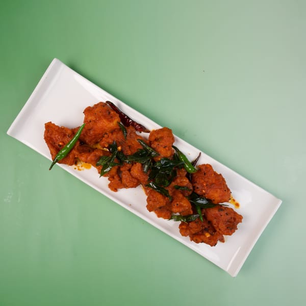 Spicy fried chicken pieces garnished with green chilies and curry leaves on a white rectangular plate.