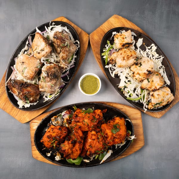 Three sizzling platters of chicken tikka, malai boti, and seekh kebab, with a green chutney.