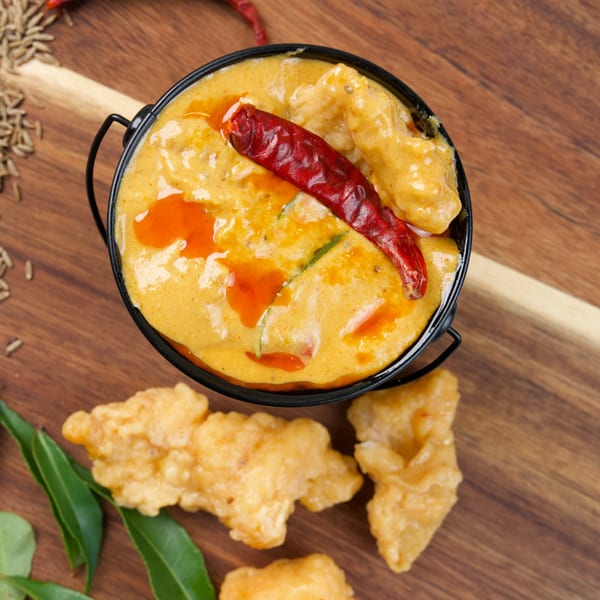 A black bowl of creamy orange curry with a red chili, surrounded by fried fritters and spices.