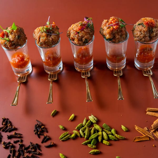 Five fried appetizers in shot glasses with sauce, garnished with herbs, on a red surface with spices.