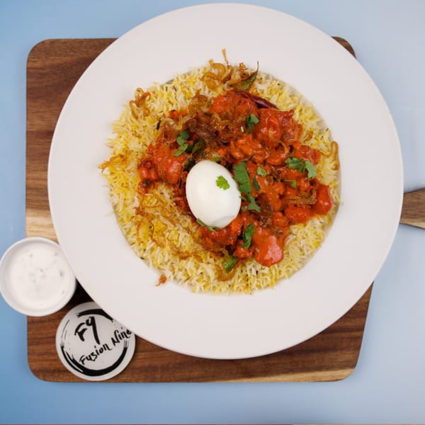 Chicken biryani with a boiled egg, fried onions, and cilantro, served with raita on a wooden board.